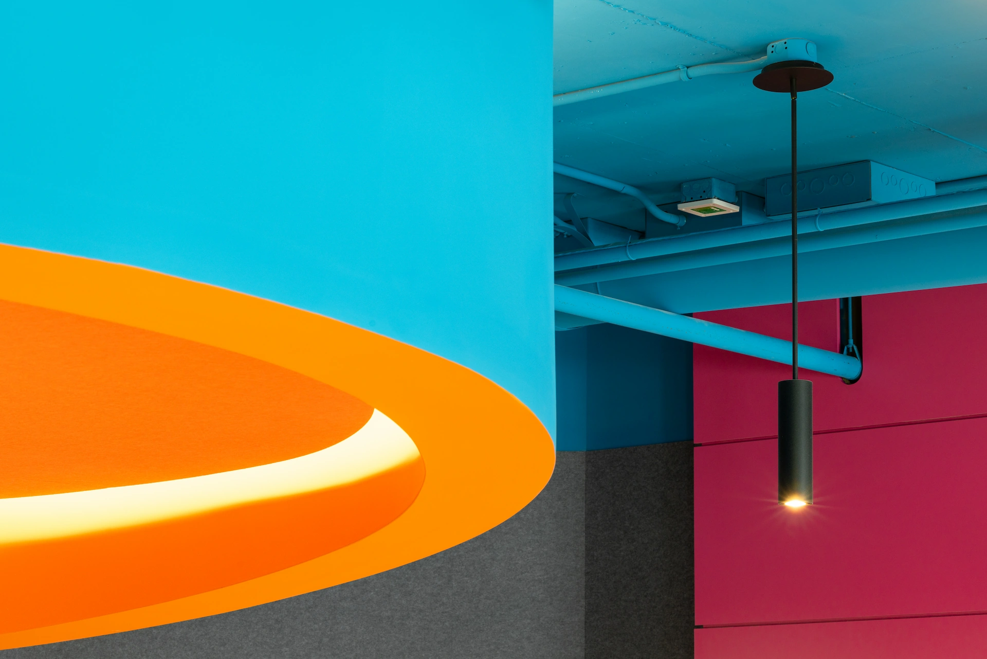 Close-up of vibrant office ceiling with bright blue exposed ductwork, curved orange acoustic feature with integrated lighting, and black cylindrical pendant light